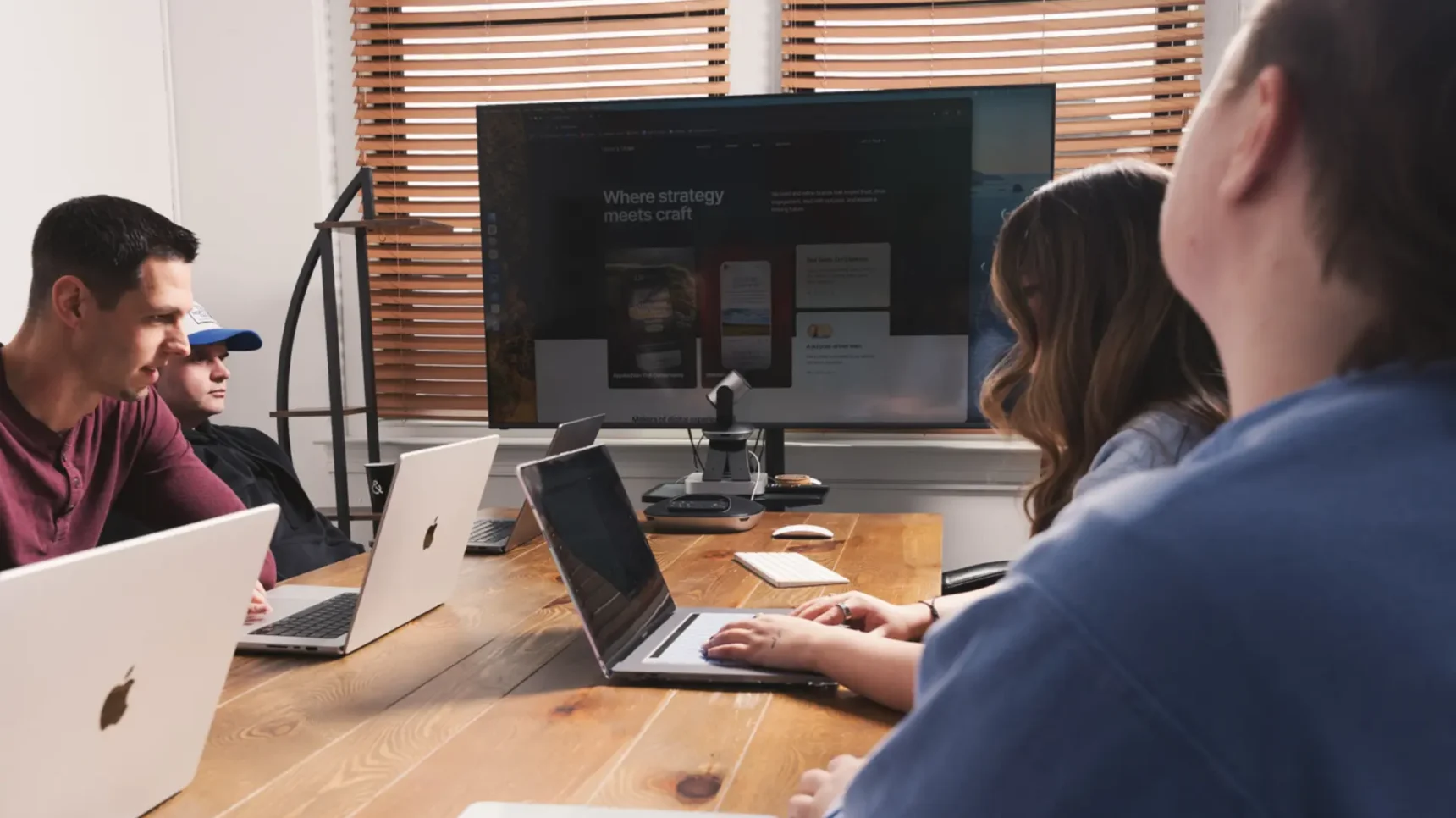 People collaborating at a desk with laptops in an office meeting, one person writing on a notepad while another views a monitor behind them, with a coffee mug on the table.