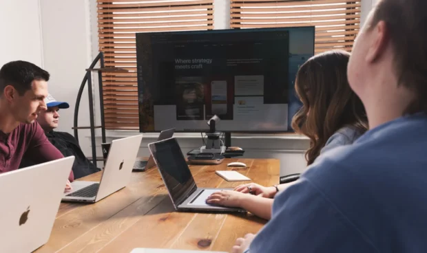 People collaborating at a desk with laptops in an office meeting, one person writing on a notepad while another views a monitor behind them, with a coffee mug on the table.