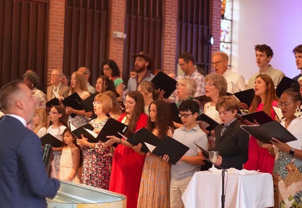 A choir in formal attire stands in front of brick walls and flowers, singing from open sheet music while a conductor in a blue suit leads with a large drum-like instrument.