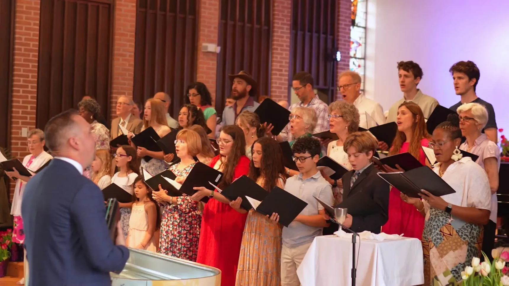 A choir in formal attire stands in front of brick walls and flowers, singing from open sheet music while a conductor in a blue suit leads with a large drum-like instrument.