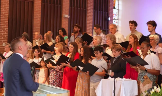A choir in formal attire stands in front of brick walls and flowers, singing from open sheet music while a conductor in a blue suit leads with a large drum-like instrument.