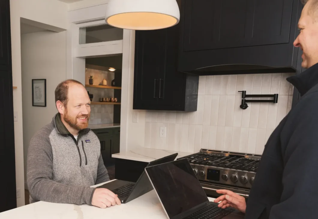 Two men stand in a modern kitchen with dark cabinets and a white countertop; one sits at a laptop while the other watches, with a wooden bowl on the counter and a stove and pendant light in view.