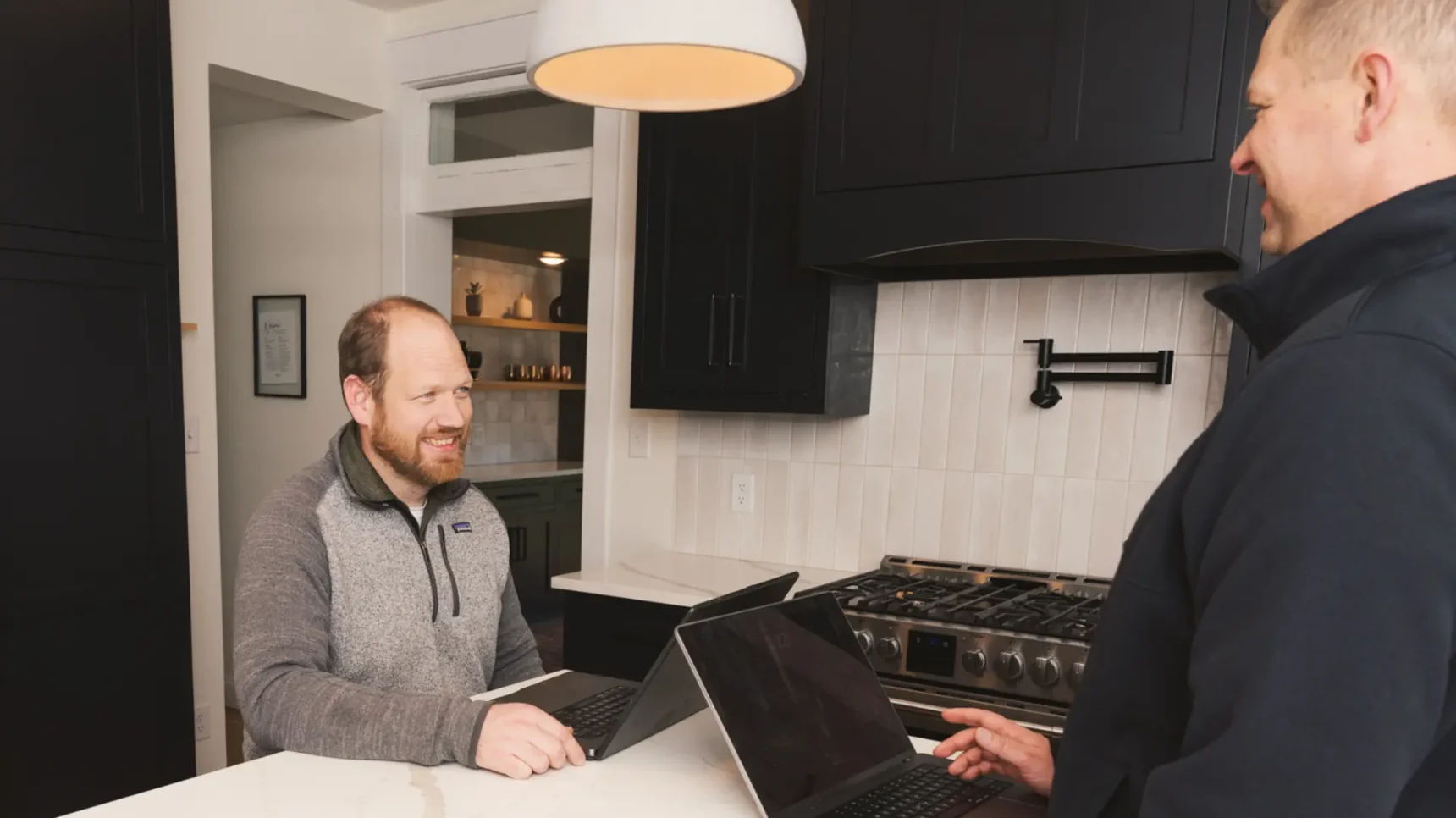 Two men stand in a modern kitchen with dark cabinets and a white countertop; one sits at a laptop while the other watches, with a wooden bowl on the counter and a stove and pendant light in view.