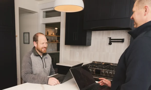 Two men stand in a modern kitchen with dark cabinets and a white countertop; one sits at a laptop while the other watches, with a wooden bowl on the counter and a stove and pendant light in view.