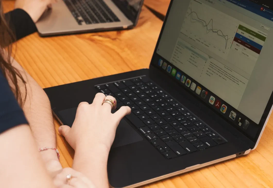 Two laptops on a wooden table while a person types with one hand and writes with the other, with a software dashboard and charts visible on the screens.