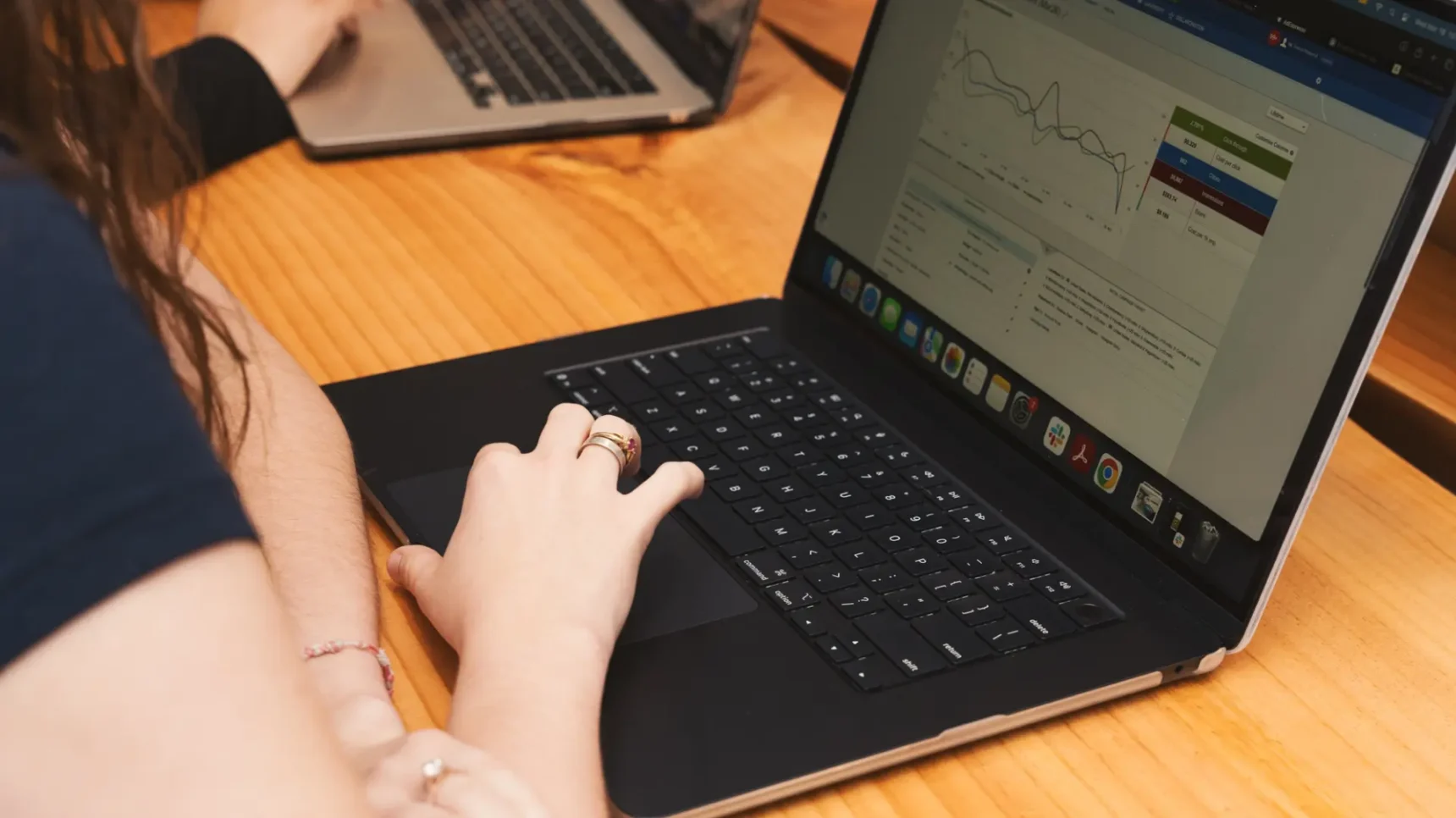 Two laptops on a wooden table while a person types with one hand and writes with the other, with a software dashboard and charts visible on the screens.