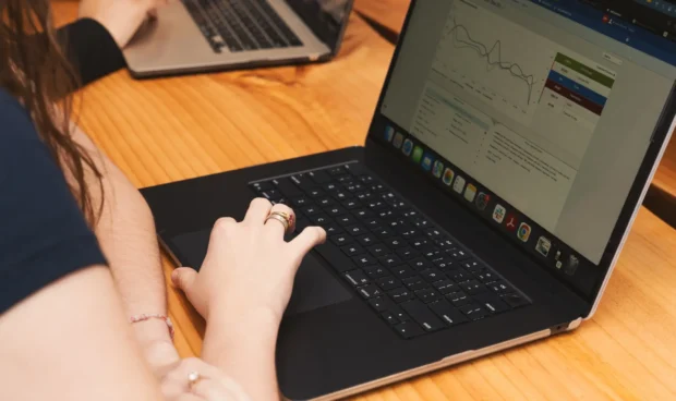 Two laptops on a wooden table while a person types with one hand and writes with the other, with a software dashboard and charts visible on the screens.